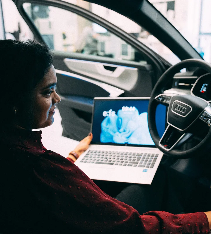 Frau sitzt auf dem Fahrersitz eines Audi-Fahrzeugs mit geöffnetem Laptop auf dem Schoß. Der Bildschirm zeigt das efs-Logo und eine blaue abstrakte Grafik. Die Fahrertür ist geöffnet, im Hintergrund ist eine Werkstatt zu erkennen.
