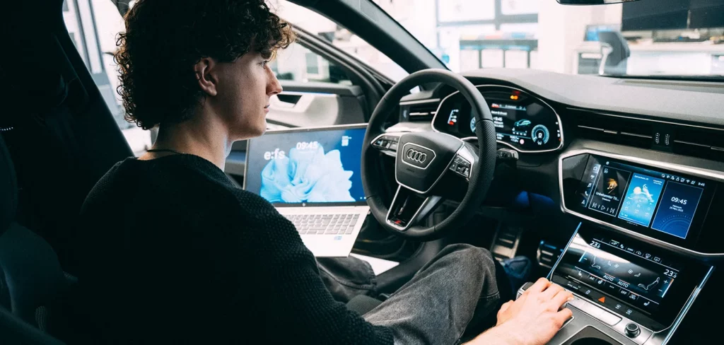 Junger Mann mit Laptop sitzt auf dem Fahrersitz eines Audi mit modernem, digitalen Cockpit. Auf dem Laptop und mehreren Fahrzeugdisplays sind Benutzeroberflächen mit Uhrzeit, Wetter und weiteren digitalen Anzeigen zu sehen.