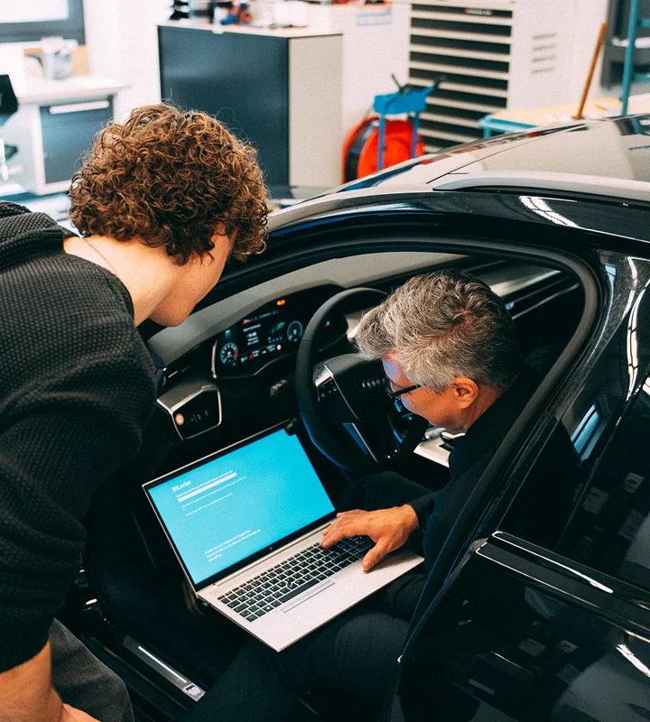 Zwei Männer arbeiten an einem Laptop im Innenraum eines Audi-Fahrzeugs. Einer sitzt auf dem Fahrersitz und bedient das Gerät, während der andere von außen über die offene Tür hereinschaut. Im Hintergrund ist eine Werkstattumgebung zu erkennen.