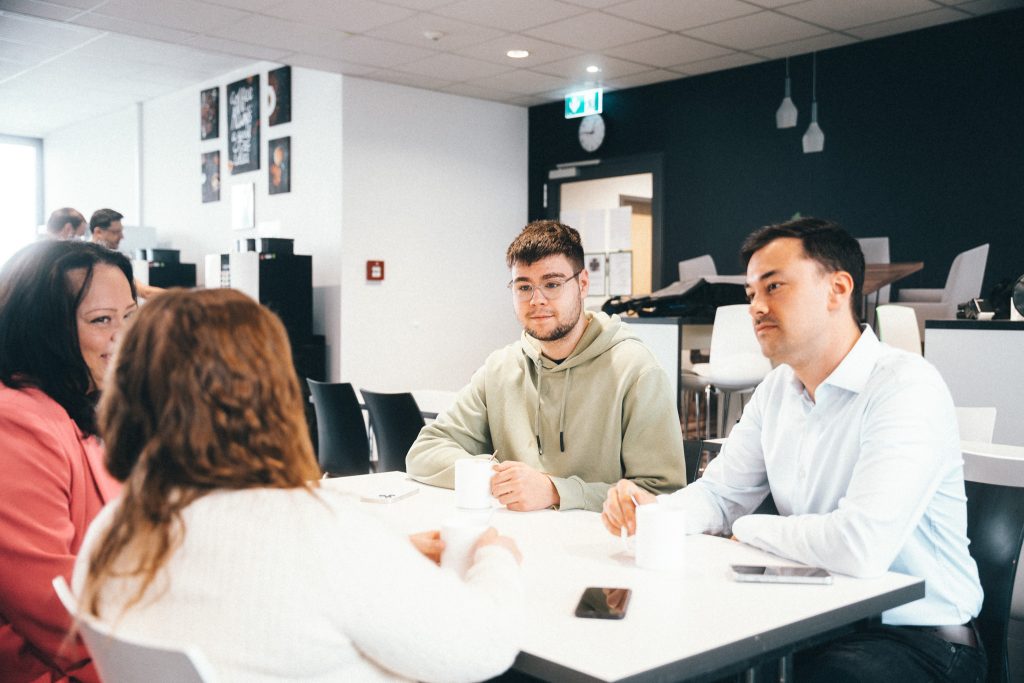 Three people are sitting at a white table with coffee cups in a modern break or communal room. In the background are a black wall, furniture, and a coffee machine. A fourth person is seen from behind.