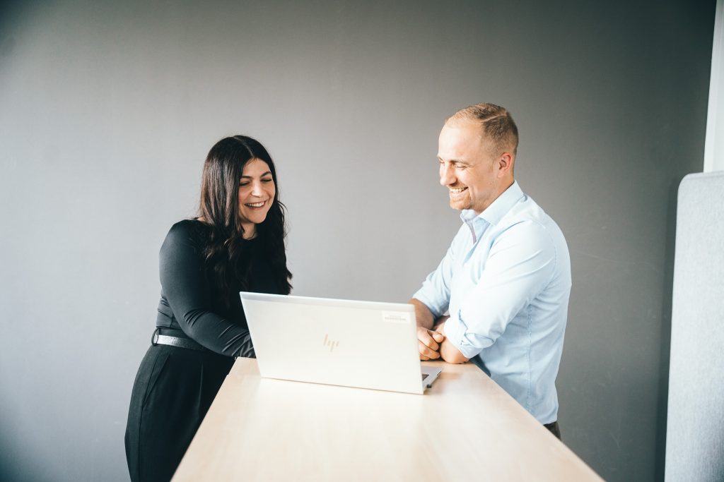 Two people are sitting next to each other at a table, looking together at an open laptop. Both are smiling. The background is neutral and gray.